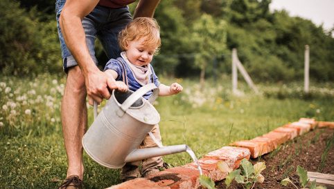 Ein Kleinkind gießt mit einer Gießkanne Gemüse in einem Garten, während ein Erwachsener ihm dabei hilft.
